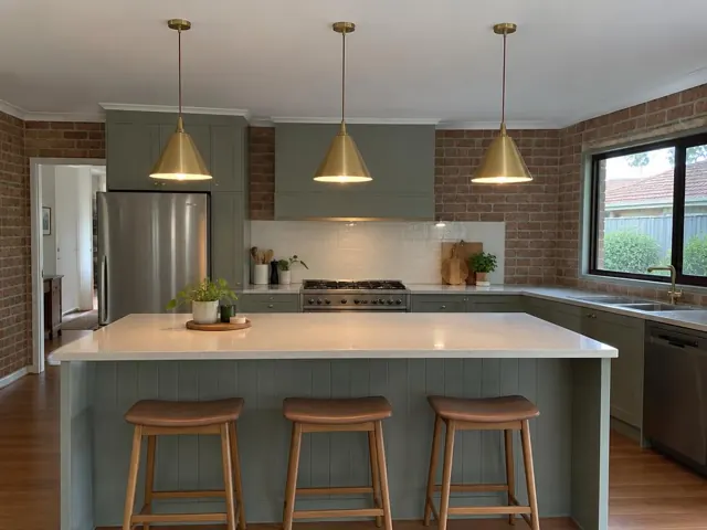 Three brass pendant lights over a kitchen island with stools, brick walls and green cabinets in Thornton